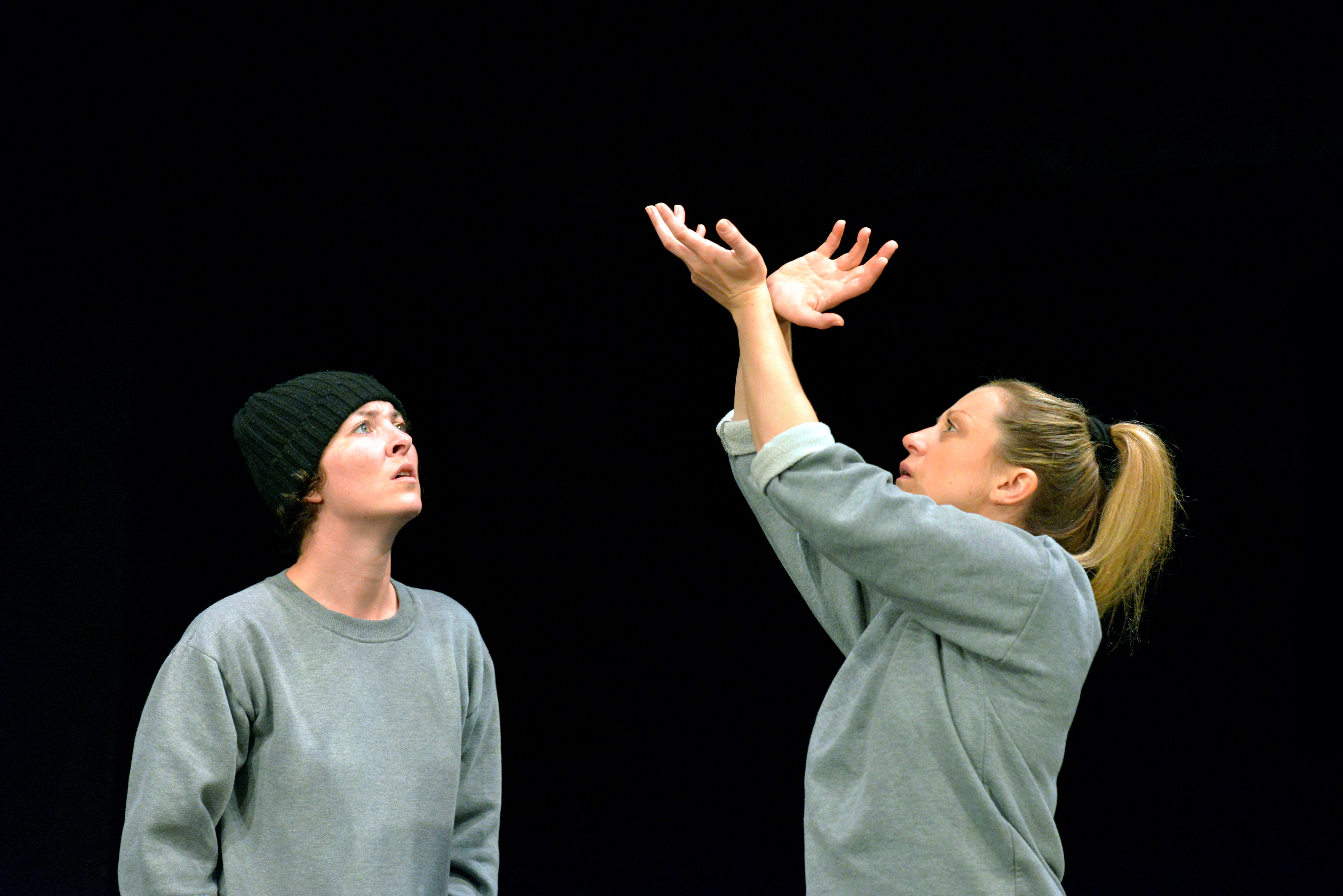Two performers in gray sweatshirts on a dark stage. One wears a black beanie and looks upward with a thoughtful expression, while the other raises their hands dramatically, palms open, appearing to gesture or reach out. Photo credit Keith Pattison